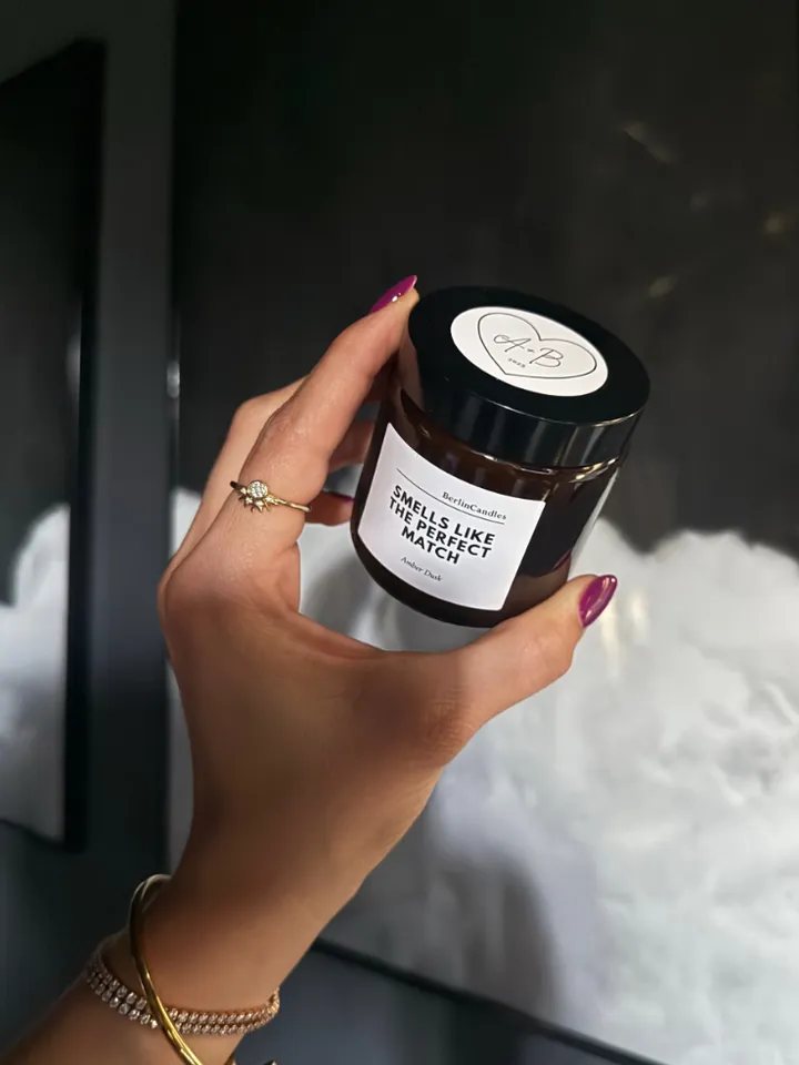 Hand with berry-painted nails and gold jewellery holding an amber glass candle; round lid label with a heart and the initials A and B, jar label reads “Smells like the perfect match”, scent Amber Dusk.