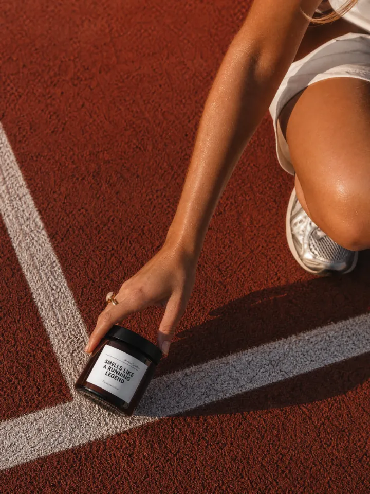 Hand placing a small amber glass candle with a black lid on the white line of a red running track; label reads “Smells like a running legend”, knee and white running shoe beside it.